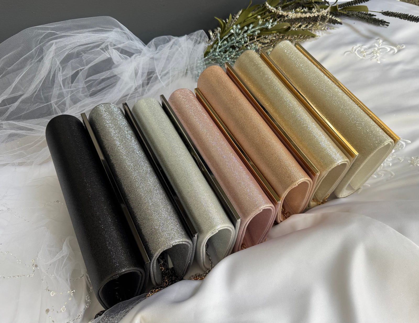 Current Lustre clutch colors lined up on white satin: black, silver, grey, pink, rose gold, gold and champagne