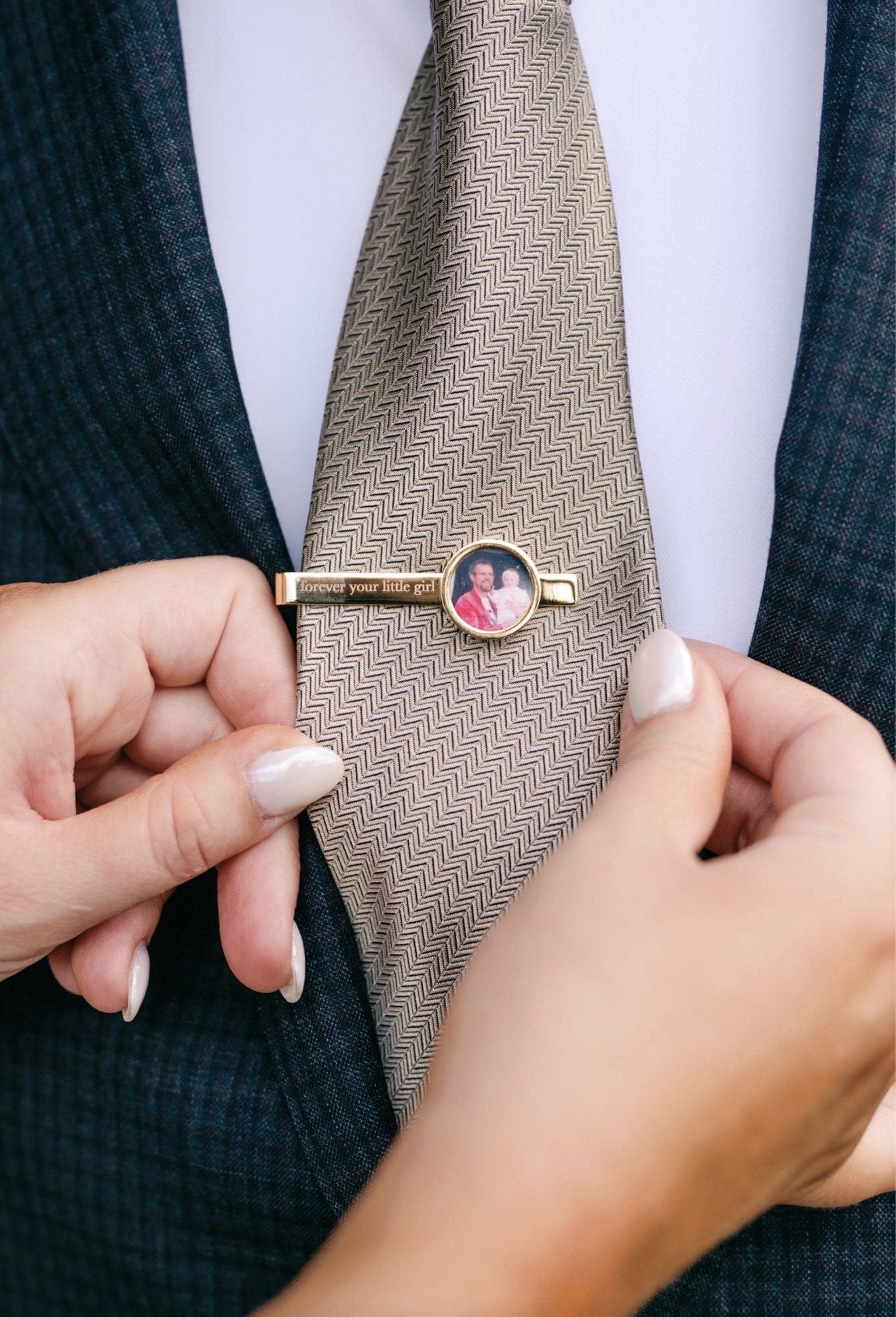 A woman’s hands adjusting a gold photo tie clip engraved “forever your little girl” on a taupe herringbone tie